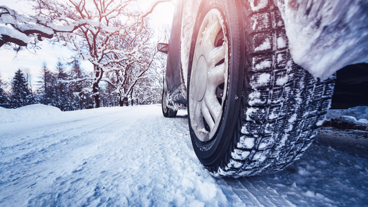 Nahaufnahme eines Autoreifens auf verschneiter Straße im Winter, umgeben von schneebedeckten Bäumen bei kaltem Sonnenlicht.