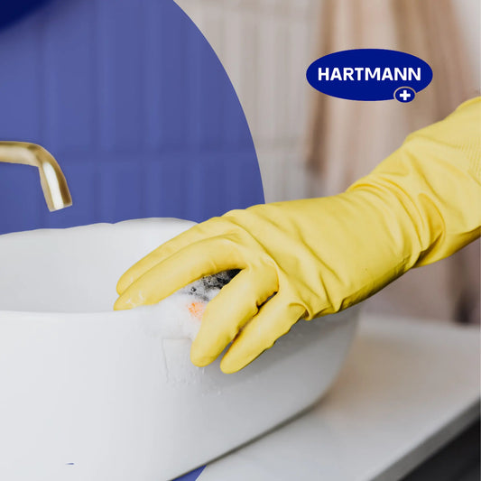 Eine Person mit einem gelben Gummihandschuh schrubbt ein weißes Waschbecken mit einem Schwamm. Auf dem Waschbecken befindet sich Seifenschaum, und ein goldener Wasserhahn ist zu sehen. Das Hartmann-Logo erscheint in der oberen rechten Ecke.