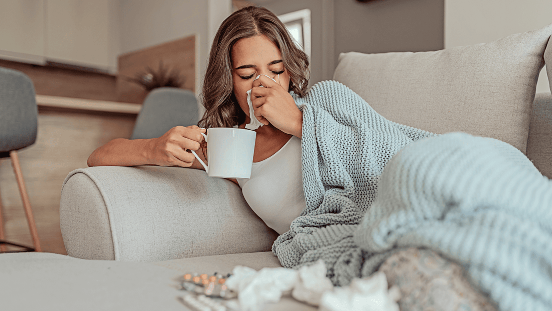Frau, die auf dem Sofa liegt und eine Tasse in der einen Hand hält, während sie sich mit der anderen die Nase putzt