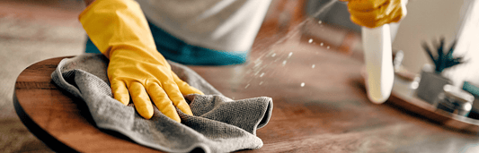 Person mit gelben Handschuhen wischt Tisch mit Tuch ab, während Reinigungsmittel daneben versprüht wird