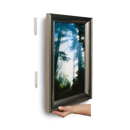 Käsi nostaa kehystetyn kuvan valkoiselta seinältä ja paljastaa kaksi 3M Deutschland GmbH:n isoa Command™-kuvankiinnitysliuskaa L, 6 sarjaa edullispakkauksessa, kuvan takana.