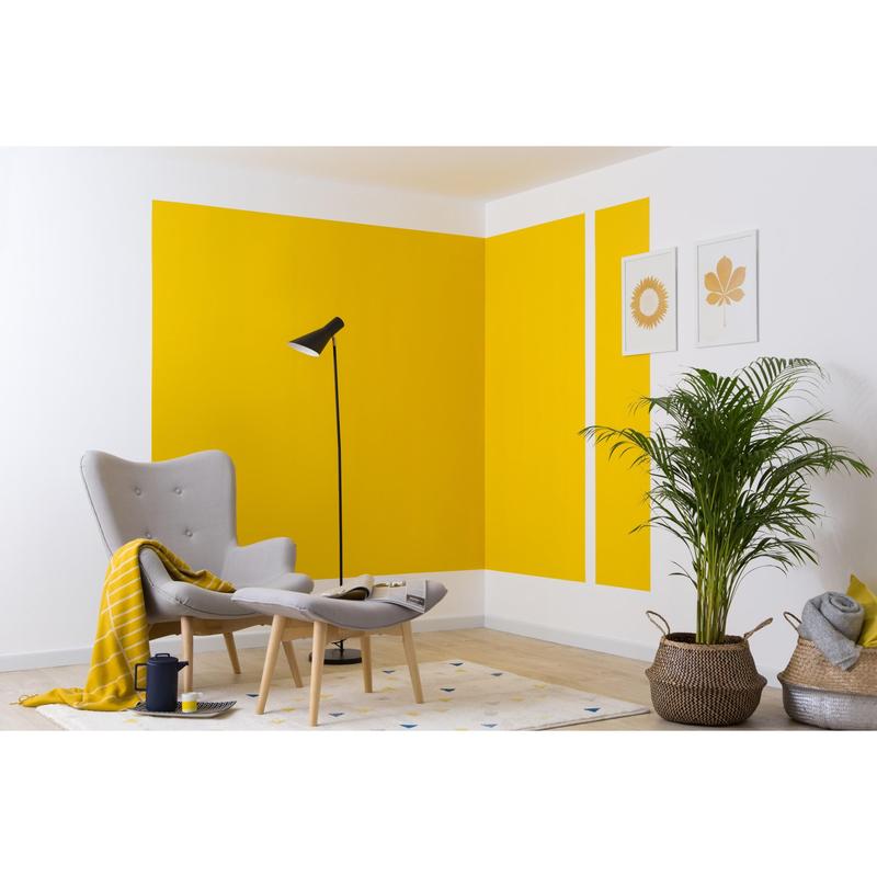 Moderni olohuone harmaalla nojatuolilla, ottomaanilla, mustalla lattiavalaisimella, keltaisella peitolla ja ruukkukasvilla. Voimakkaat keltaiset geometriset muodot seinällä on luotu 3M Deutschland GmbH:n ScotchBlue™ maalarinteipillä teräville linjoille.