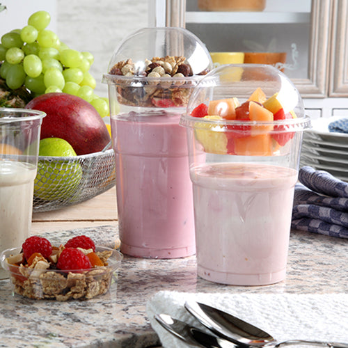 Kaksi jogurttiparfaitia muovikupeissa, jokainen suljettuna Starpak 50 Dom-kannella „Hurricane“ PET pyöreä kirkas Ø 9,5 cm, seisoo keittiötasolla - yksi myslillä ja pähkinöillä, toinen hedelmillä. Taustalla näkyy tuoreita marjoja ja pinottuja lautasia.