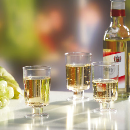 Kolme Starpak-valkoviinilasia (PS, 0,1l, kirkas) on täytetty ja ne seisovat valkoviinipullon ja vihreiden rypäleiden vieressä valkoisella pinnalla epätarkalla taustalla.