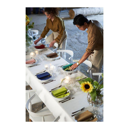 Zwei Personen arrangieren bunte Servietten auf einem langen, mit Gläsern und Besteck gedeckten Tisch. Eine Dunicel-Tischdeckenrolle (1,18 m x 10 m) der Duni GmbH deckt den Tisch ab und verleiht ihm den Charme großer Veranstaltungen im einladenden Innenbereich.