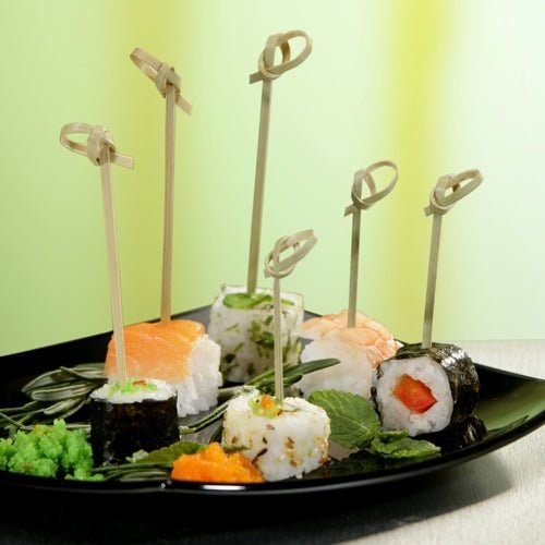 Musta lautanen, jossa on erilaisia susheja, jokainen pala on pistetty PAPSTAR 40 Fingerfood - piikillä „pure“ PAPSTAR GmbH:lta ja koristeltu yrteillä ja kalamädillä, esitetään elegantisti taustalla, jossa on vihreä värigradientti.