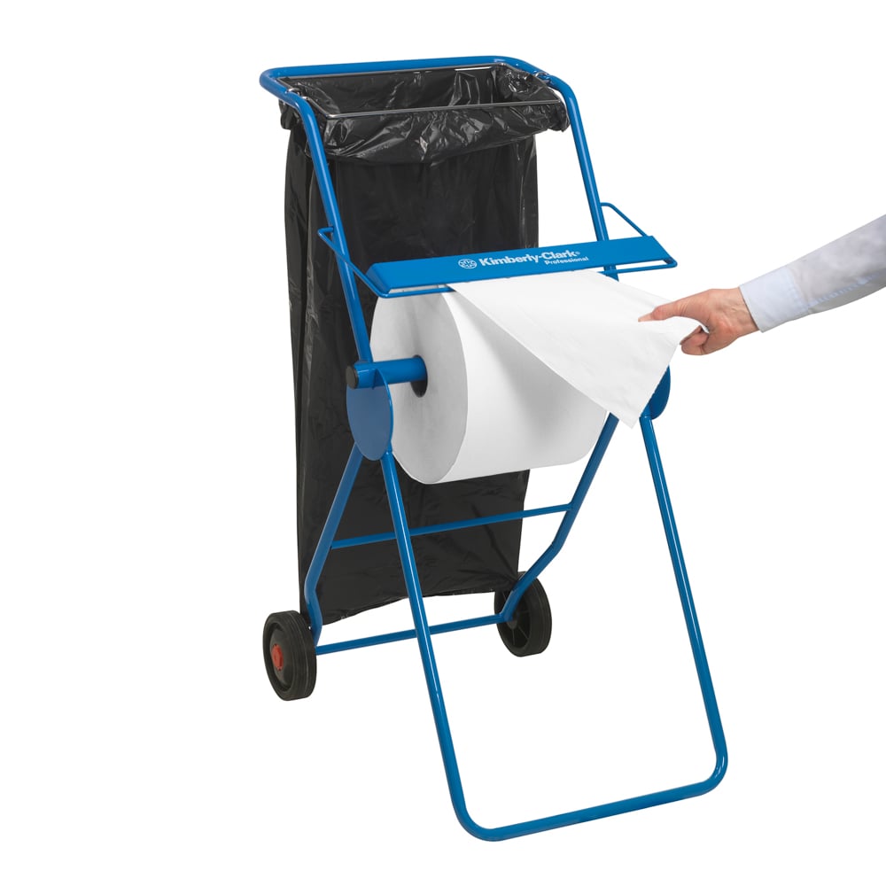 Sininen paperipyyheannostelija, jossa on suuri valkoinen Scott® ESSENTIAL™ lääkärirulla (6 x 200 arkkia) Kimberly-Clark GmbH:lta korostaa hygieniaa; musta roskapussi on lähellä, kun henkilön käsi vetää arkin rullasta.