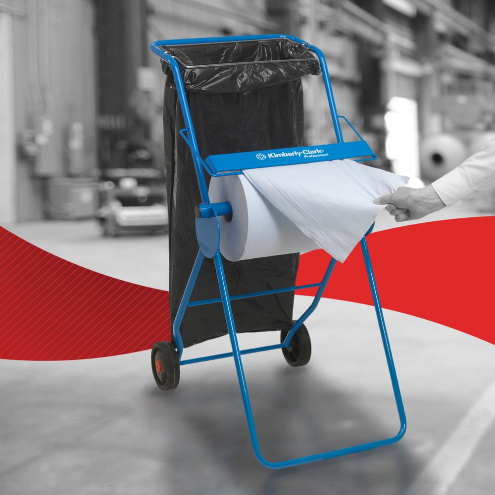 Ein blauer Kimberly-Clark-Bodenständer hält ein WypAll® L20 Großrolle Papierwischtuch für Instandhaltungsarbeiten und einen schwarzen Müllsack. Eine Hand zieht ein Tuch - ideal für industrielle Wartungsarbeiten in Innenräumen.