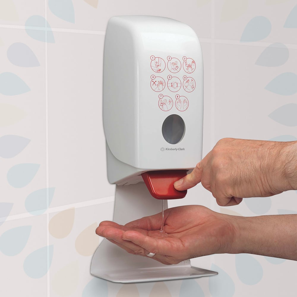 Henkilö painaa punaista vipua seinään kiinnitetyssä Kimberly-Clark GmbH:n Kleenex® Extra Gel -käsidesiannostelijassa ja annostelee kirkasta käsidesiä kädelleen. Etupuolella on kuvallinen ohje, ja taustalla on kuvioituja kaakeleita.