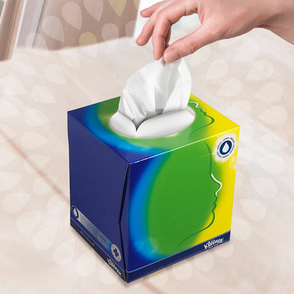 Käsi vetää nenäliinan Kleenex® kosmetiikkapyyhkeistä - kuutio-laatikko / valkoinen Kimberly-Clark GmbH:lta, jossa on sininen, vihreä ja keltainen design ja valkoinen soikea aukko. Tyylitelty profiili viittaa kasvojen hoitoon pehmeästi sumentuneen taustan edessä.