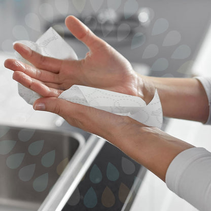 Henkilö pyyhkii käsiään valkoisilla Kleenex® Rollenhandtücher E-roll Slimroll™ paperipyyhkeillä (1 kerros, 6x135m) Kimberly-Clark GmbH:lta pesualtaan yllä, ja vesipisaroita on lisätty kuvaan.
