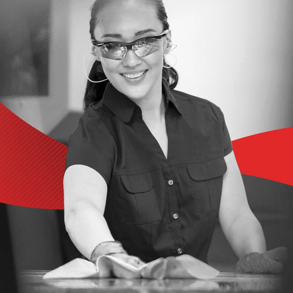 Hansikkaisiin ja silmälaseihin pukeutunut nainen hymyilee puhdistaessaan pintaa Kimberly-Clark GmbH:n WypAll® X60 General Clean - BRAG™ Box -pyyhkeellä (41,80 × 28,30 cm); taustalla näkyy punainen graafinen suunnittelu.