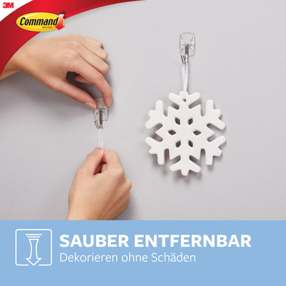 Eine Hand befestigt einen Command™ Drahthaken, klein, transparent, mit transparenten Strips von 3M Deutschland GmbH an der Wand und hängt eine weiße Schneeflocke auf. Text: "Sauber entfernbar. Dekorieren ohne Schäden.