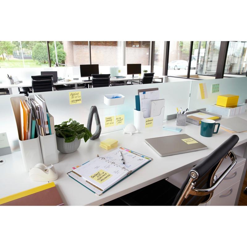 Moderni toimistotyöpaikka, jossa on työpöytä kansioiden, kannettavan tietokoneen, suunnittelijan ja 3M Deutschland GmbH:n keltaisten Post-it® Notesien (76 mm x 127 mm, 100 arkkia/lohko, 12 lohkoa/pakkaus) kanssa, suuret ikkunat ja tyhjät tuolit taustalla.