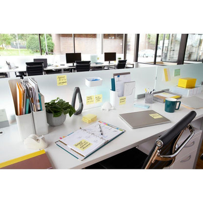 Moderni toimistotyöpaikka, jossa on työpöytä kansioiden, kannettavan tietokoneen, suunnittelijan ja 3M Deutschland GmbH:n keltaisten Post-it® Notesien (76 mm x 127 mm, 100 arkkia/lohko, 12 lohkoa/pakkaus) kanssa, suuret ikkunat ja tyhjät tuolit taustalla.