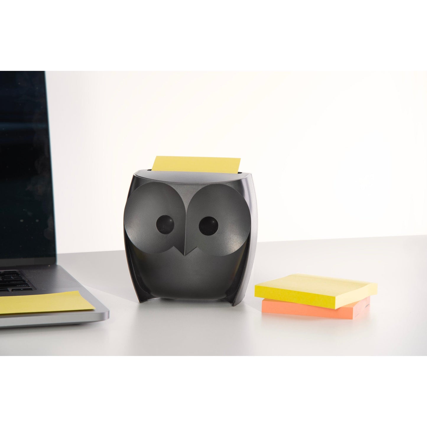Ein 3M Post-it® Z-Notes Dispenser in Eulenform, schwarz, hält gelbe Super Sticky Z-Notes. Daneben liegen ein geschlossener Laptop und eine Packung mit gelben und pinken Z-Notes (2 Blöcke à 45 Blatt) auf einem weißen Schreibtisch.