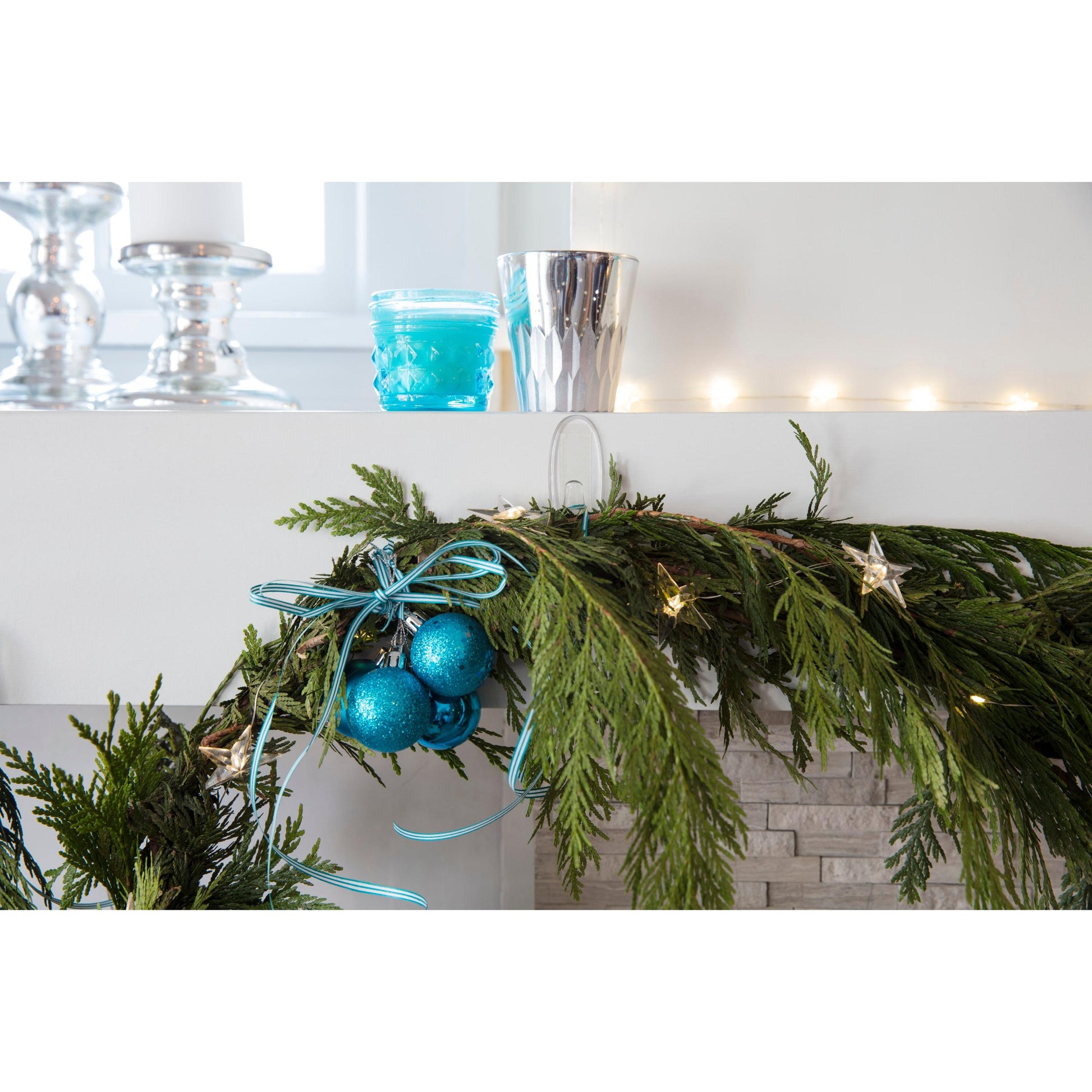 Ein Kaminsims ist mit grüner Tannengirlande, blauen Bändern, warmen Lichterketten, Kerzen und einem blauen Glas und einem silbernen Becher geschmückt - alles sicher aufgehängt mit den transparenten Command™ Haken M der 3M Deutschland GmbH.