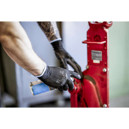 Henkilö, jolla on NITRAS-nailonkäsineet, mustat, PU-päällystetyt AS Arbeitsschutz GmbH:ltä, puhdistaa tai voitelee sinisellä suihkepullolla punaista metallista konenosaa. Näkyvissä ovat vain käsivarret ja kädet.