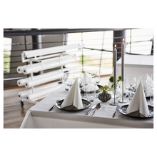 Auf einem hübsch gedeckten Esstisch steht eine frische, weiße Duni Dunicel®-Tischdeckenrolle der Duni GmbH, die eine stoffähnliche Haptik bietet. Dreieckig gefaltete Servietten auf dunklen Tellern begleiten Weingläser. Hohe Kerzen und kleine Blumenarrangements dienen als Tischdekoration, im Hintergrund sind gerollte Leinwände zu sehen.