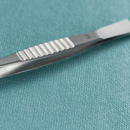 Lähikuva Paul Hartmann AG:n Hartmannin suorasta 9 cm lastapinsetistä, steriili (40 kappaleen pakkaus), joka sijaitsee metallisen kirurgisen veitsen vieressä turkoosilla, teksturoidulla lääketieteellisellä liinalla.