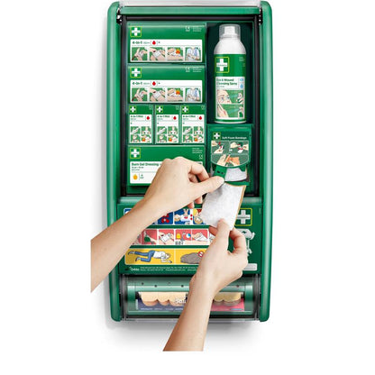 Henkilö pitää sidettä Cederroth ensiapuaseman edessä Orkla Wound Care AB:ltä, joka on varustettu siteillä, palovammageelillä, suolaliuoksella ja sidoksilla. Kuvitetut ohjeet ovat nähtävissä sisäpuolella olevissa hyllyissä.