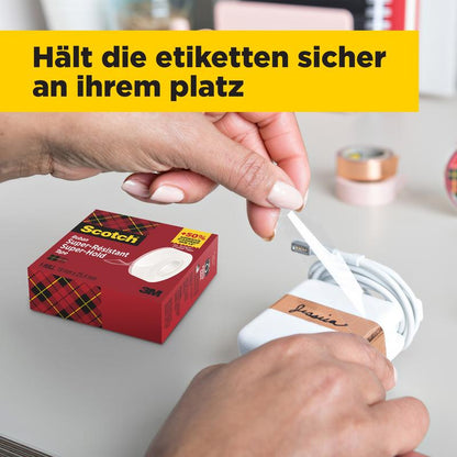 Henkilö kietoo valkoista etikettiä mustalla tekstillä valkoisen laturin ympärille, kun pöydällä on laatikko Scotch® Super-Hold Secure Tape -teippiä, 1 rulla, 19 mm x 25,4 m 3M Deutschland GmbH:ltä. Taustalla on epätarkkoja toimistotarvikkeita ja keltainen Saksan lippu.