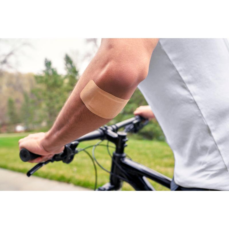 Henkilö, joka käyttää valkoista paitaa ja ajaa pyörällä ulkona, on kiinnittänyt Nexcare™ Ultra Stretch MAXI Comfort Flexible -laastarin (50 mm x 101 mm) 3M Deutschland GmbH:lta paljaaseen olkavarteen; taustalla näkyy vihreää ruohoa ja puita.