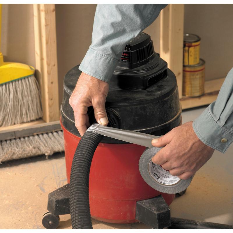 Henkilö käärii Scotch® Universal Teippiä 2904 (50 m x 48 mm) 3M Deutschland GmbH:lta pölynimurin letkun ympärille, joka on kiinnitetty punaiseen työpajaimuriin. Taustalla näkyy luuta ja maalipurkkeja.