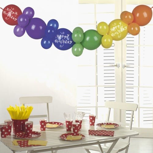 Valkoinen pöytä, jossa on punapilkulliset juhla-astiat, keltaiset välineet ja kupit, on kruunattu PAPSTAR Ballonkoristeella 2 m "Hyvää Syntymäpäivää" PAPSTAR GmbH:ltä - juhlava syntymäpäiväkoriste, joka loistaa auringonvalossa valkoisten ikkunaluukkujen läpi.