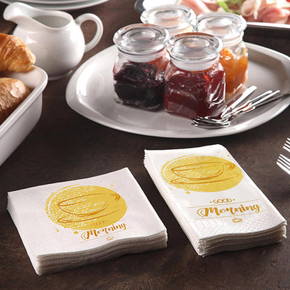 Aamiaispöytä croissanteilla, kannulla ja 200 PAPSTAR lautasliinalla (3-kerroksinen 1/4-taitos, 24x24 cm) PAPSTAR GmbH:lta, joissa lukee „Hyvää huomenta“, lisäksi hillopurkit ja kolme haarukkaa mukavaa aamiaista varten.