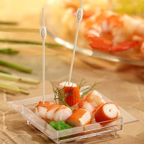 Neliönmuotoisella muovialustalla, jossa on rapujäljitelmää, katkarapuja, vihreää mätiä ja koristeita, on kaksi PAPSTAR 40 Fingerfood - piikkiä „pure“ PAPSTAR GmbH:lta, taustalla näkyy epätarkasti vastaava merenelävälautanen.