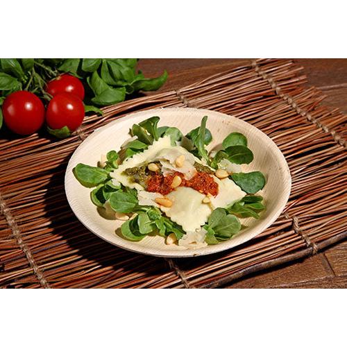 Raviolilautanen tomaattikastikkeella, pinjansiemenillä ja juustolla lehtivihannesten päällä tarjoillaan PAPSTAR 25 lautasella, palmunlehti „pure“ pyöreä PAPSTAR GmbH:lta, joka lepää bambu-pöytäliinalla kirsikkatomaattien ja lehtivihannesten taustalla.