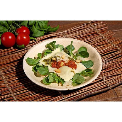 Raviolilautanen tomaattikastikkeella, pinjansiemenillä ja juustolla lehtivihannesten päällä tarjoillaan PAPSTAR 25 lautasella, palmunlehti „pure“ pyöreä PAPSTAR GmbH:lta, joka lepää bambu-pöytäliinalla kirsikkatomaattien ja lehtivihannesten taustalla.
