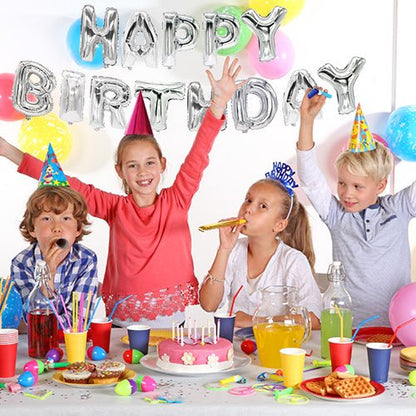 Neljä lasta juhlahatuissa juhlivat syntymäpäivää koristellun pöydän ääressä, jossa on kakku ja juhlatavaroita. Taustalla seinää koristavat "Happy Birthday" -folioilmapallosetti PAPSTAR GmbH:ltä hopeisena ja värikkäinä ilmapalloina.