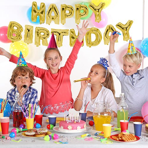 Neljä lasta juhlahatuissa istuvat juhlallisesti katetun pöydän ääressä kakkujen ja juomien kera. Seinällä folioilmapallosetti "Happy Birthday" PAPSTAR GmbH:ltä tuo kultaista hohtoa syntymäpäiväjuhlaan.