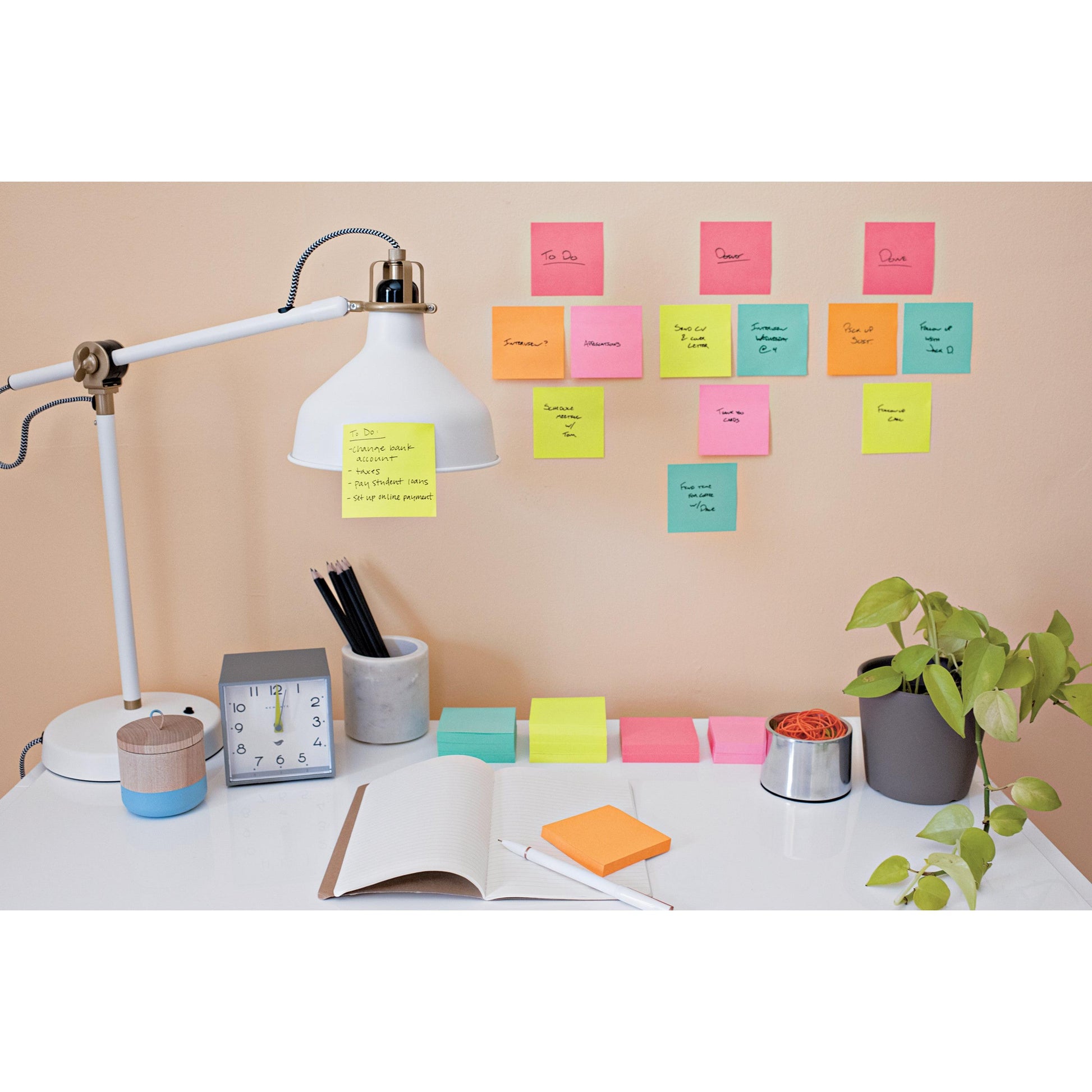 Ein aufgeräumter Schreibtisch mit weißer Lampe, Uhr, Stiften, Pflanze, aufgeschlagenem Notizbuch und ultrahellen 3M Post-it® Notes Würfeln (76x76 mm, 450 Blatt) der 3M Deutschland GmbH in leuchtenden Farben an der Wand.