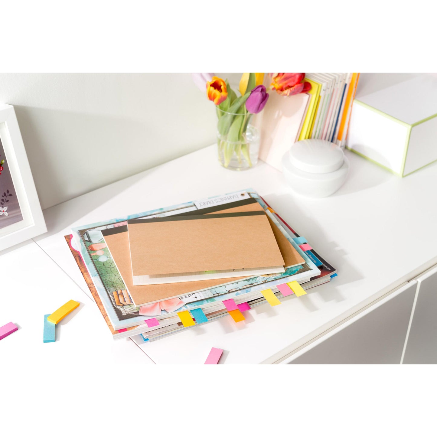 Ein Stapel Zeitschriften und ein Notizbuch mit bunten 3M Deutschland GmbH Post-it® Page Marker (15x50 mm, 5 Blöcke/Packung) liegt auf einem weißen Schreibtisch, in dessen Nähe lose Zettel verstreut sind. Im Hintergrund stehen eine Vase mit Tulpen und einige Bücher.