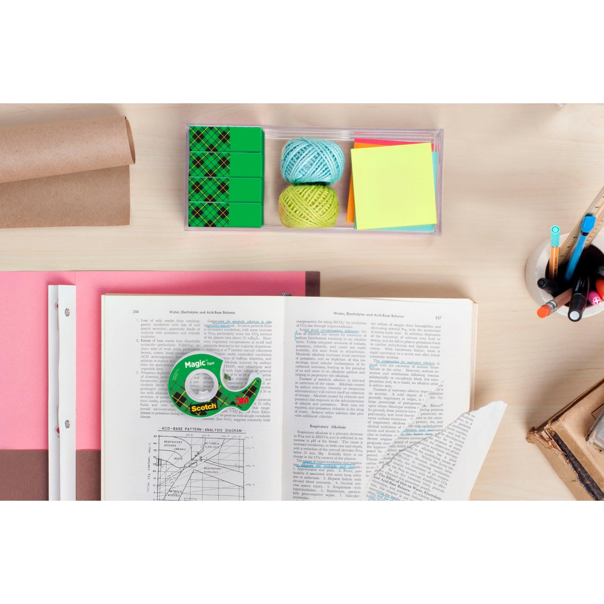 Ein Schreibtisch mit aufgeschlagenen Büchern, einer zerrissenen Seite, farbigen Haftnotizen, Radiergummis, Stiften, Bleistiften, Papier, Garnkugeln, einer Pappröhre und Scotch® Magic™ Unsichtbares Klebeband Vorteilspack von 3M Deutschland GmbH.