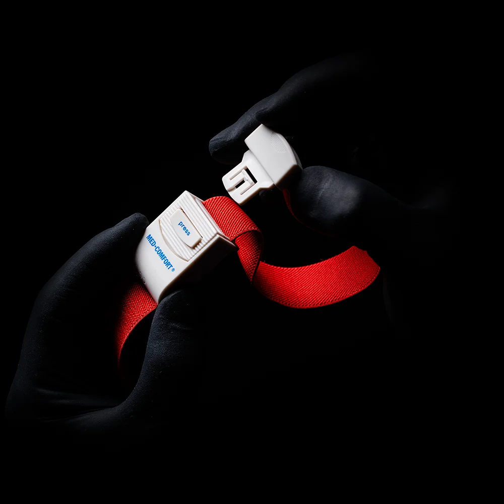 Käsineet kädet, jotka muistuttavat AMPri MED-COMFORT -verenkierron kiristintä, tarttuvat kirkkaan punaisen turvavyön solkeen varjoisalla taustalla. Soljessa näkyvät sana 'Blue' ja kirjain 'B'.