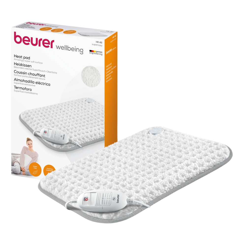 Pakkauksen vieressä on Beurer HK 42 Super-Cosy -lämpötyyny Beurer GmbH:n hyvinvointimerkillä. Valkoinen, harmaareunainen tyyny on varustettu kaukosäätimellä. Pakkaus näyttää lisäksi kuvan käytössä olevasta lämpötyynystä ja korostaa pikakuumennustoimintoa.