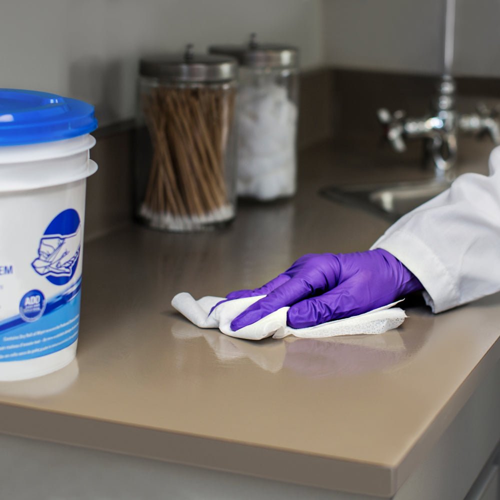 Henkilö violeteissa hanskoissa pyyhkii Kimberly-Clark GmbH:n WypAll® Wettask™ -pyyhkeillä (6 rullaa x 90 arkkia) beigeä työtasoa pesualtaan lähellä, samalla kun taustalla näkyy purkkeja vanupuikkojen ja -pallojen kanssa.