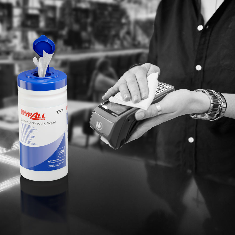 Jemand benutzt ein WypAll® Flächendesinfektionstuch (Kimberly-Clark GmbH) aus einer offenen Spenderwanne zur Desinfektion eines Kartenterminals. Der weiße Spendereimer (200 Tücher) steht auf der Theke, der Hintergrund ist unscharf.
