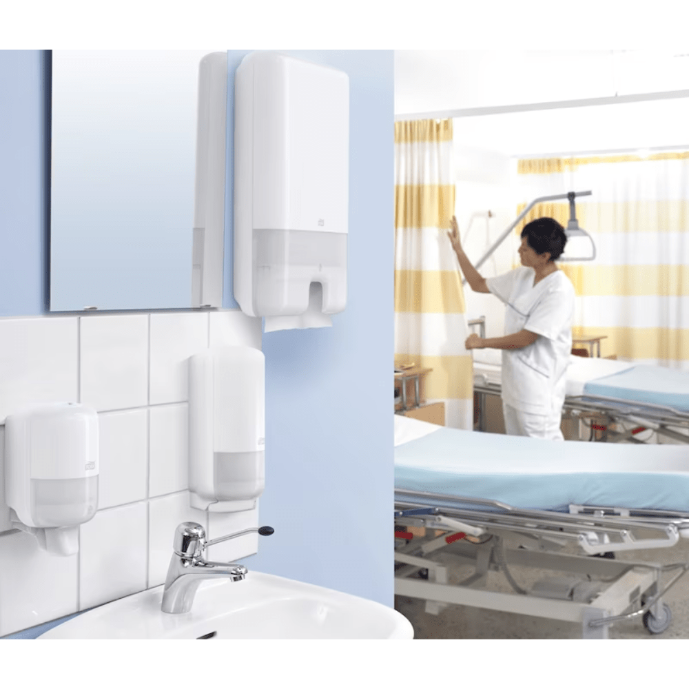 Sairaalahuoneessa seisoo lääkäri valkoisessa univormussa sängyn vieressä. Etualalla on pesuallas, jossa on Tork Xpress® 552000 -annostelija Multifold-käsipyyhkeille H2 TORK - Essity Professional Hygiene Germany GmbH:lta. Huone on varustettu lääketieteellisillä laitteilla ja keltaisilla raidallisilla verhoilla.