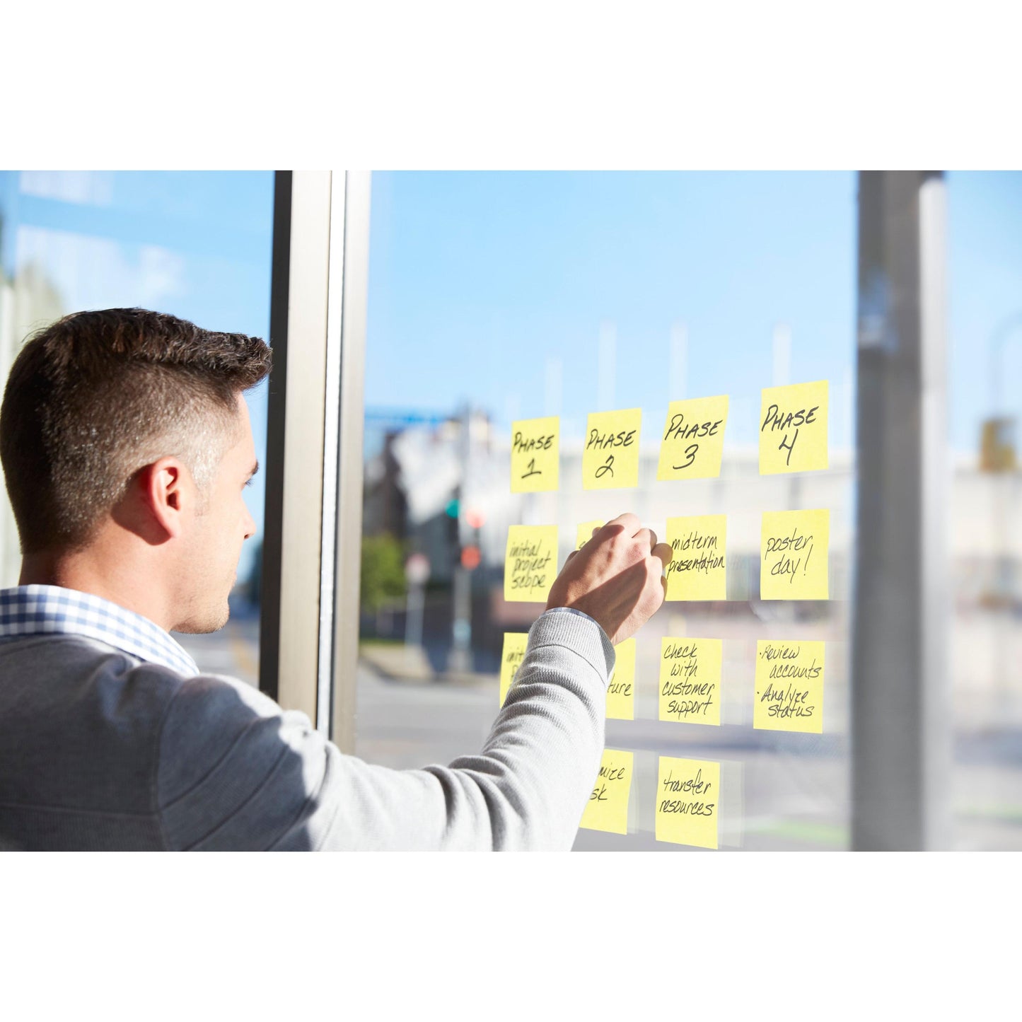 Ein Mann steht an einem Fenster und ordnet 3M Deutschland GmbH Post-it® Super Sticky Notes, Gelb, 51 mm x 76 mm, auf denen Projektphasen und Aufgaben mit schwarzem Marker geschrieben sind. Die Zettel sind zur besseren Visualisierung in Spalten auf dem Glas angeordnet.