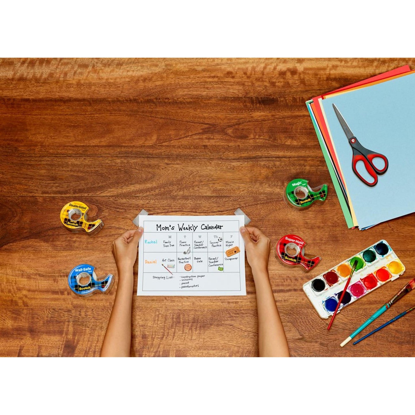 Eine Person hält einen "Mom's Weekly Calendar" auf einem Holztisch mit vier Klebebandabrollern, farbigem Papier, einem Aquarellset mit Pinsel und einer Scotch™ Präzisionsschere rot von 3M Deutschland GmbH.