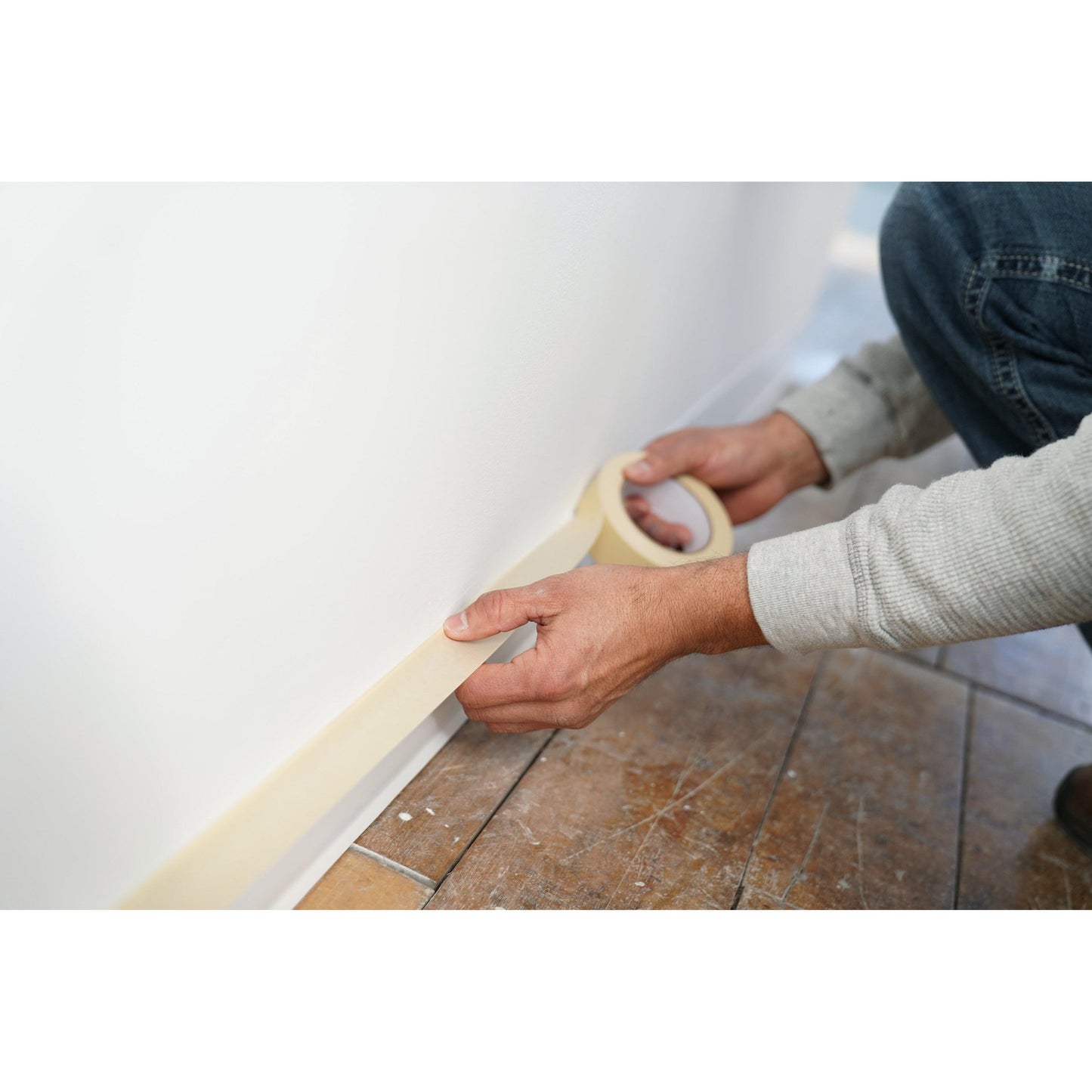 Eine Person verwendet Scotch® Classic Abdeckband (24 mm x 50 m, 1 Rolle/Packung, 70% PEFC) der 3M Deutschland GmbH, um bei Maler- und Renovierungsarbeiten den Sockel einer weißen Wand über einem Fliesenboden abzudecken.