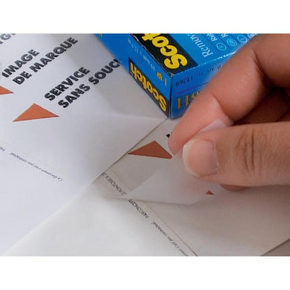 Käsi irrottaa kolmiomaisen tarran arkista, jossa on teksti "IMAGE DE MARQUE SERVICE SANS SOUCI", samalla kun taustalla on 3M Deutschland GmbH:n Scotch® näkymätön teippi, irrotettava, 19 mm x 33 m, 1 rulla | 1 kappale.