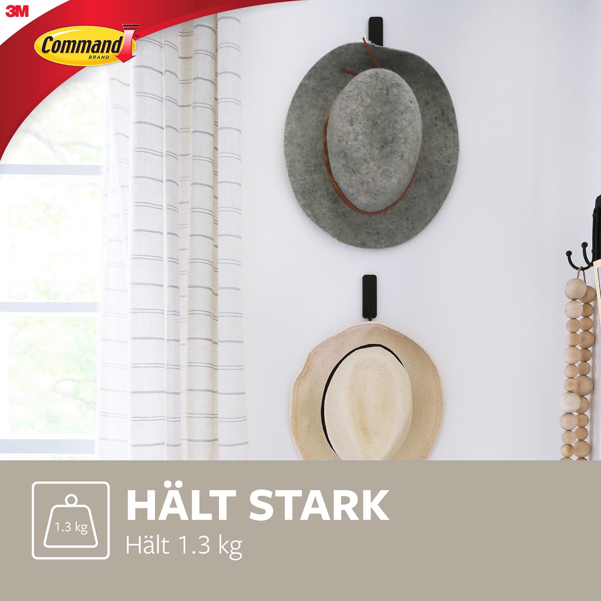 Zwei Hüte, ein grauer und ein beiger, hängen mit einem mittelgroßen Command™ Mattschwarzer Haken (17034B) von 3M Deutschland GmbH an einer weißen Wand. Ein Fenster mit weißen Vorhängen und einer Perlendekoration ist ebenfalls zu sehen. Es erscheinen deutscher Text und das Command-Logo.