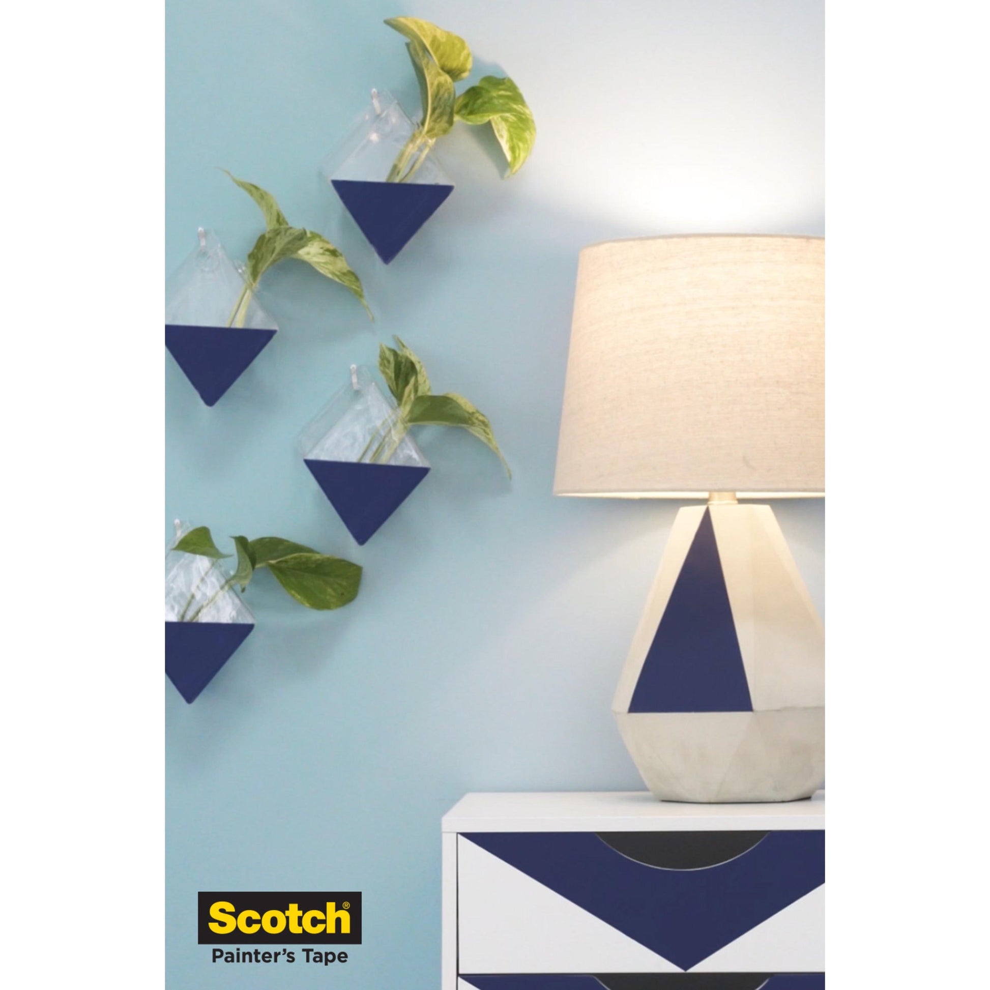 Eine geometrische Lampe und eine Kommode mit blauen und weißen Mustern stehen neben einer blauen Wand, die mit drei dreieckigen Pflanzgefäßen verziert ist. In der linken unteren Ecke ist ein Logo der 3M Deutschland GmbH Scotch® Malerabdeckband für empfindliche Oberflächen zu sehen.