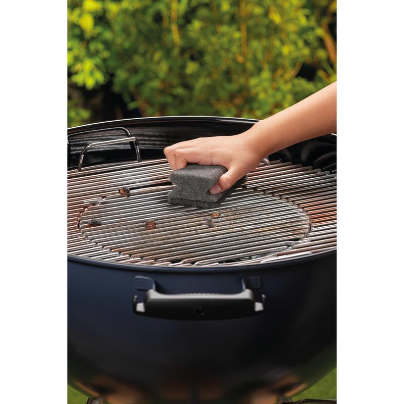 Käsi puhdistaa pyöreän hiiligrillin metalliritilän Scotch-Brite® Grillgriffschwammilla (1 kappale per pakkaus) 3M Deutschland GmbH:lta. Vihreät lehdet ovat epätarkkoina taustalla.
